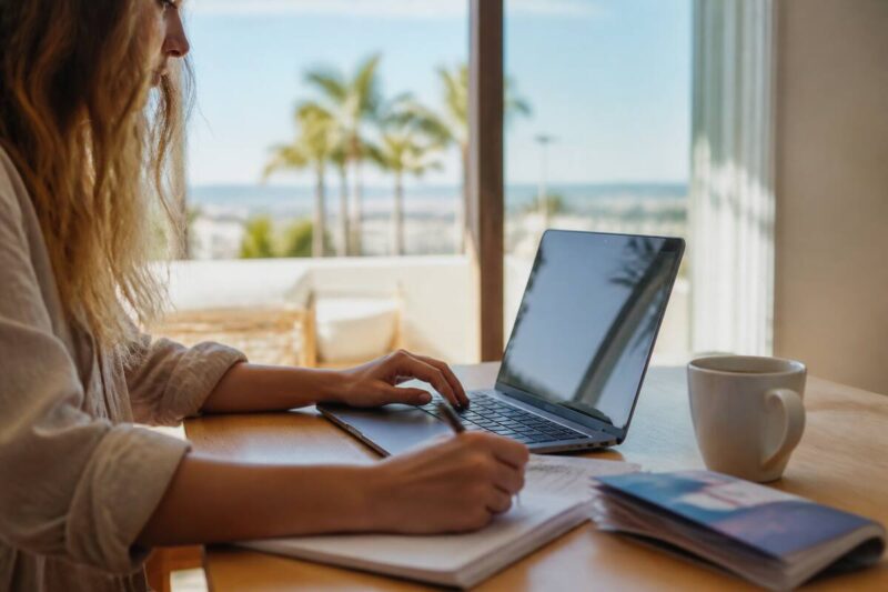 Iemand vergelijkt vakanties op een laptop met reisnotities op tafel